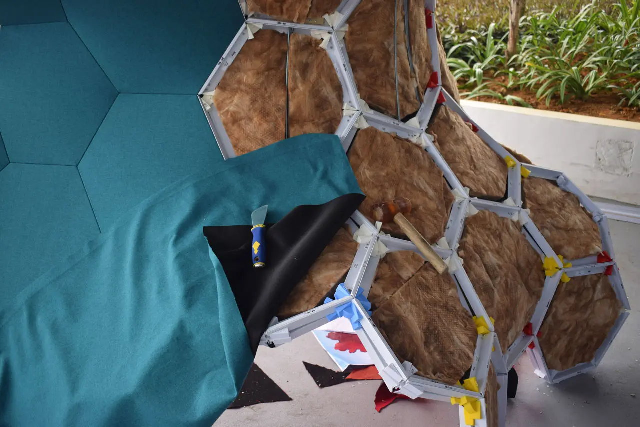 A partially assembled geodesic structure with hexagonal panels, some covered in teal fabric and others exposing insulation and wood, was featured at SingaPlural 2019. A utility knife and fabric rest on the structure.