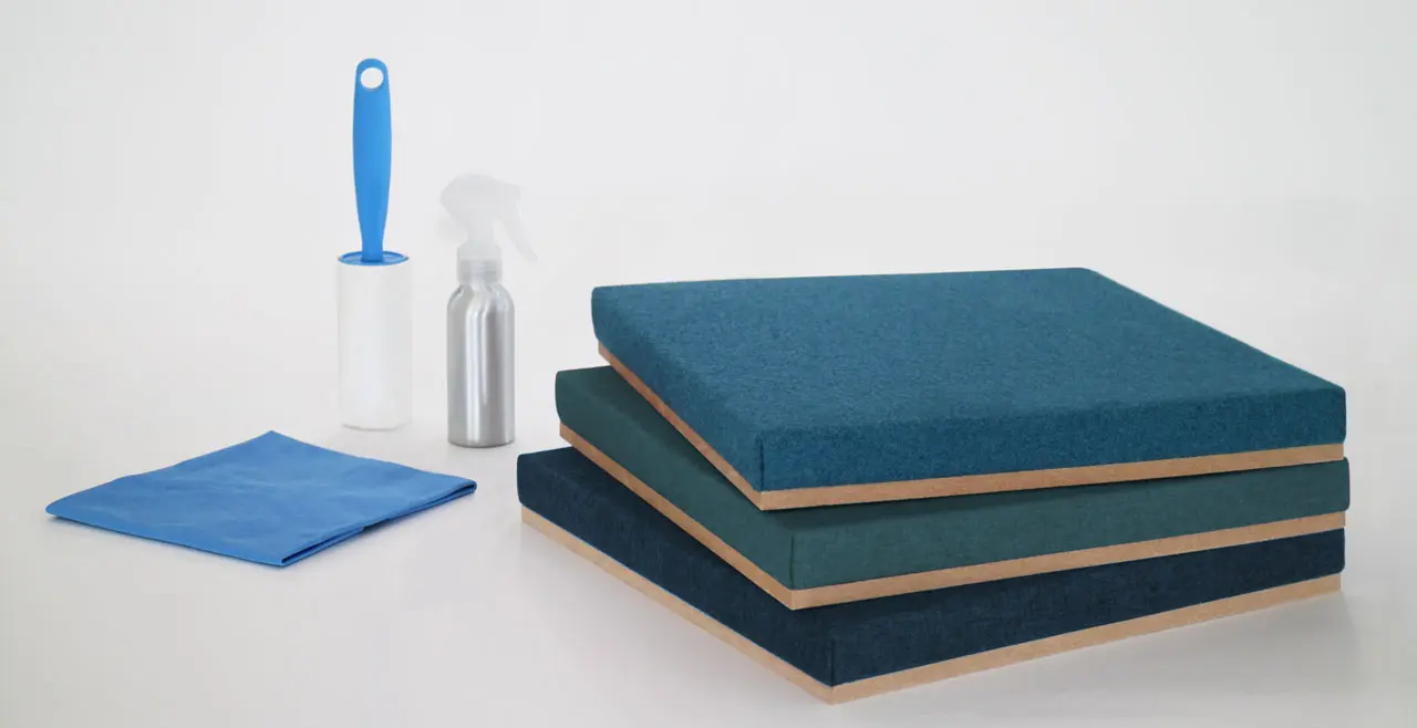 Three blue rectangular fabric cushions are stacked on the right. On the left, a blue lint roller, silver spray bottle, and folded blue cleaning cloth sit against a white background—perfect for maintaining your Fabrix™ Acoustic System Warranty.