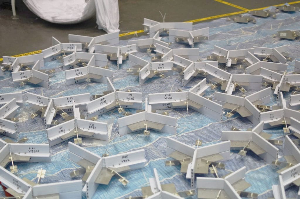 Numerous metal corner brackets with labels are scattered across a blue tarp on a workshop floor. Yellow caution tape outlines the area.