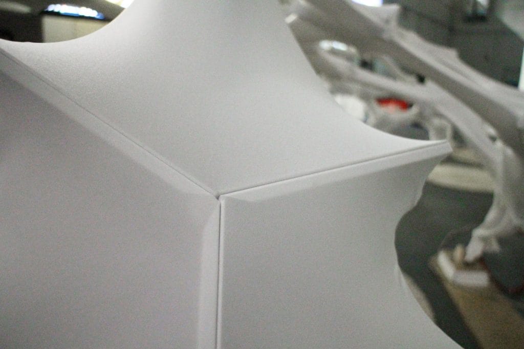 Close-up of a geometric white structure with sharp edges and smooth surfaces, forming a three-way corner. The background is blurred, showing other similar white forms and a gray setting.