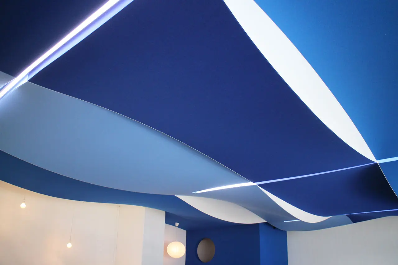 Curved blue and white ceiling panels create a wave-like pattern, with integrated linear lights. Round wall lights hang on the white wall below the modern ceiling design.