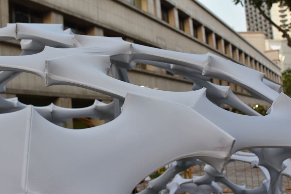 Close-up of a modern, geometric white outdoor sculpture made of interconnected, stretched fabric panels, set in front of a multi-story concrete building with windows.