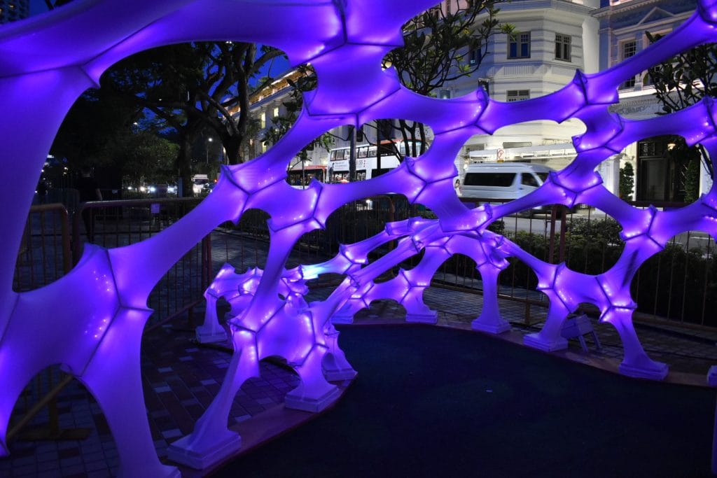 A glowing, purple, web-like art installation is displayed outdoors at night, with trees, railings, and a bright white building visible in the background. The structure is illuminated from within, creating a vibrant scene.