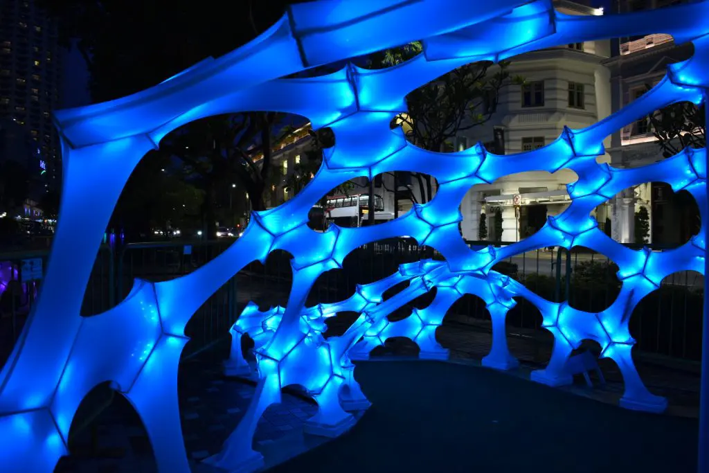 A glowing blue art installation with a web-like pattern is displayed outdoors at night, illuminating the surrounding area with bright blue light. Buildings and trees are visible in the background.