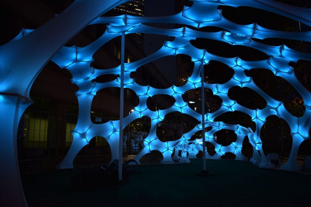 A glowing blue geometric canopy structure illuminated at night, made of interconnected hexagonal shapes, with city buildings and lights visible in the background.