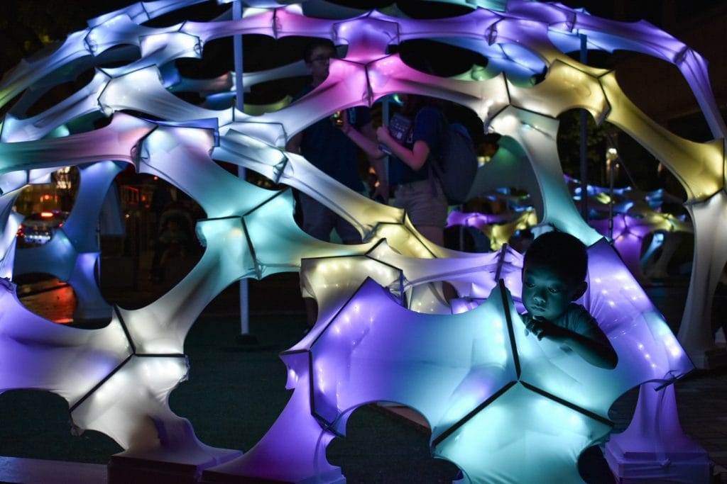 A young child stands inside a glowing, colorful geometric structure lit by multicolored lights at night, with people in the background exploring the illuminated installation.