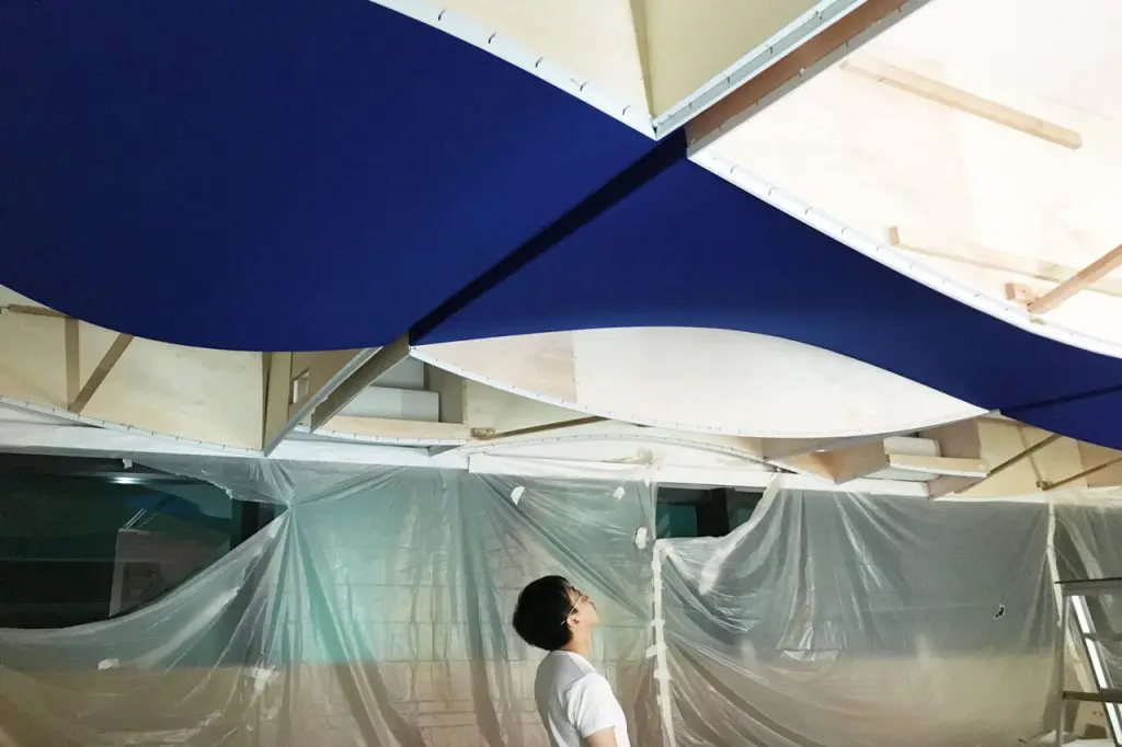 A person looks up at a ceiling installation featuring large, wavy blue panels and exposed wooden beams, with plastic sheeting covering walls and objects in the background.