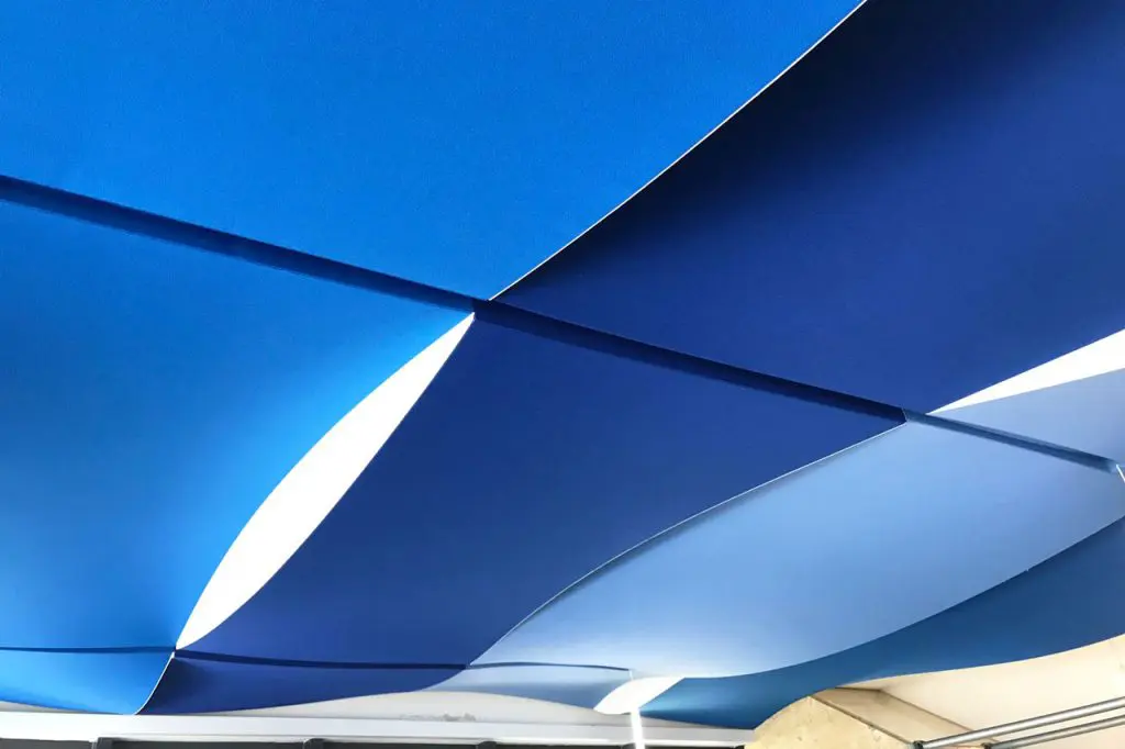 Geometric ceiling panels in shades of blue and white overlap and intersect, creating a modern, abstract pattern. The design features sharp angles and contrasting colors, giving the ceiling a dynamic, three-dimensional appearance.
