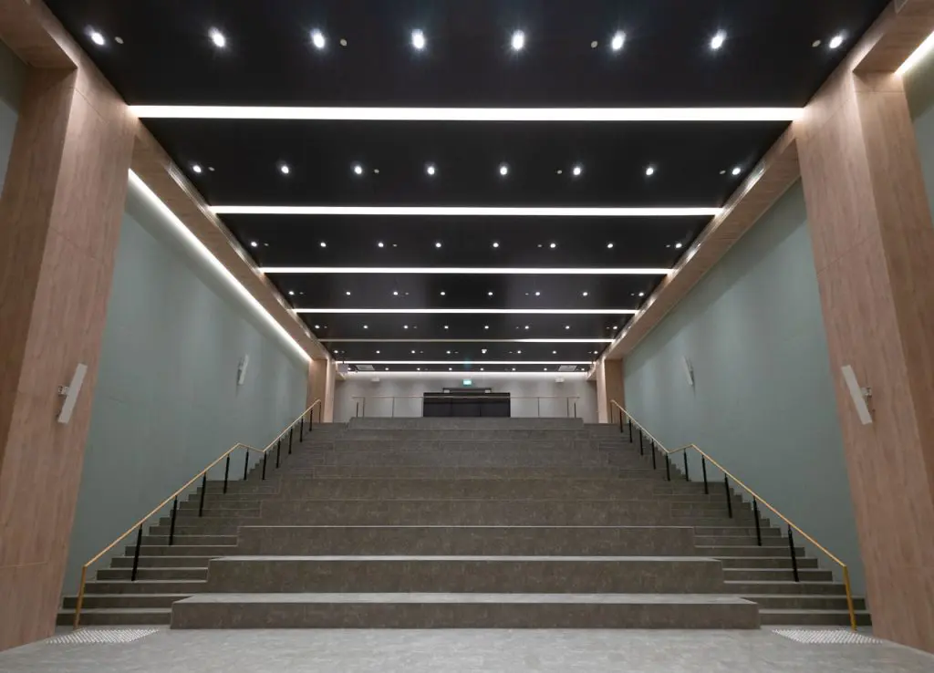 Wide staircase with broad steps and railings on both sides, leading up to a modern platform in a well-lit hall with wood paneling, a black ceiling featuring rows of recessed lights, and an Auditorium Fabric System Installation.