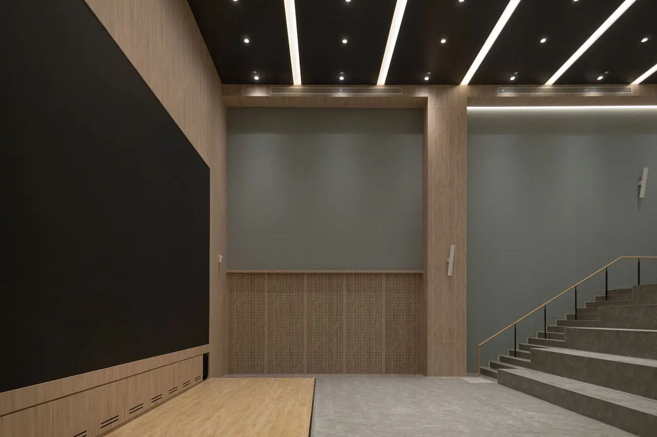 A modern, minimalist auditorium with Auditorium Fabric System Installation, featuring a large black screen, wood-paneled walls, stepped seating, and recessed ceiling lights. The space showcases clean lines and neutral colors.