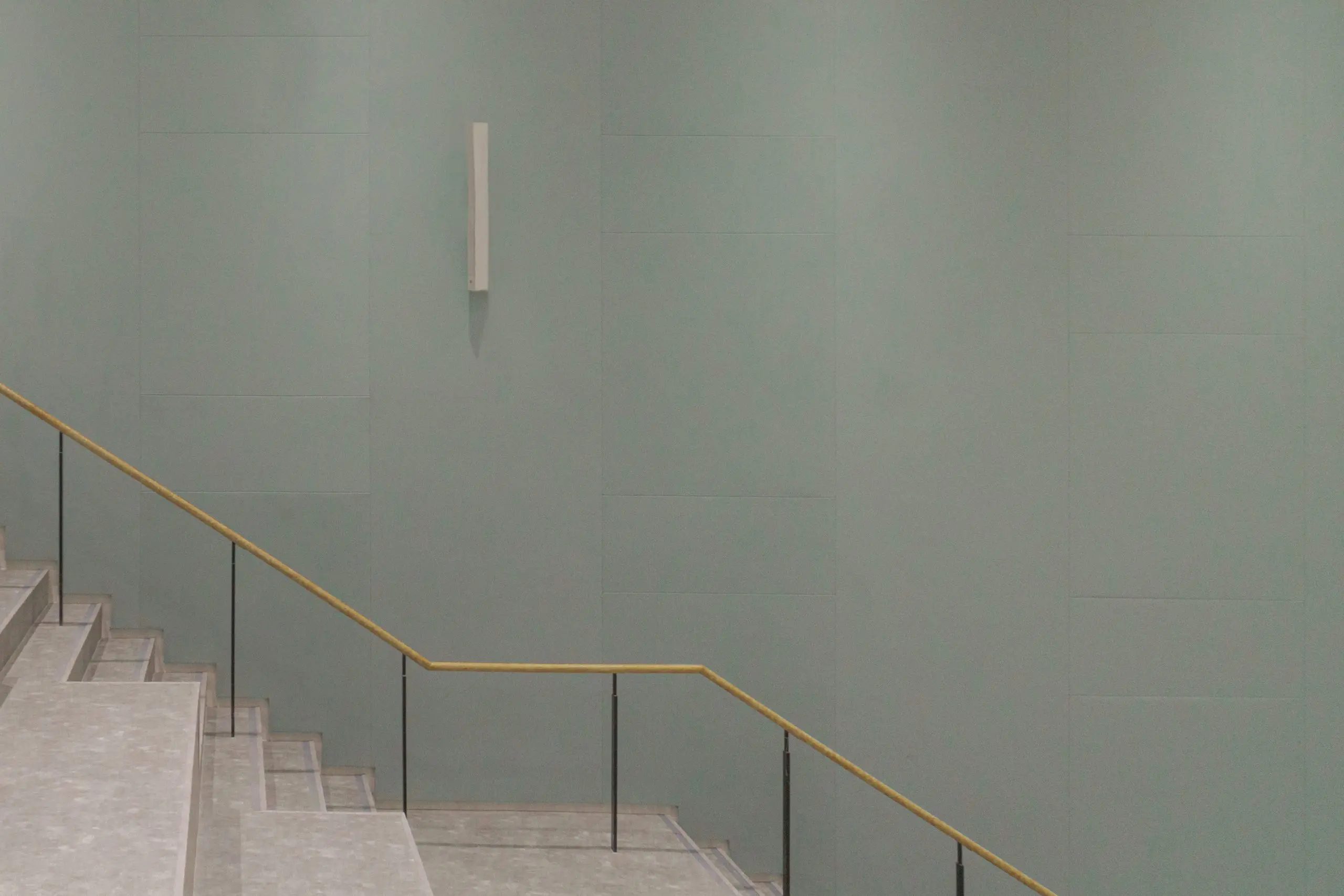 Wide staircase with light gray steps and a thin wooden handrail ascends beside a large, smooth, pale green wall. A minimalist vertical light fixture is mounted above the stairs, complementing the clean lines of the Auditorium Fabric System Installation.