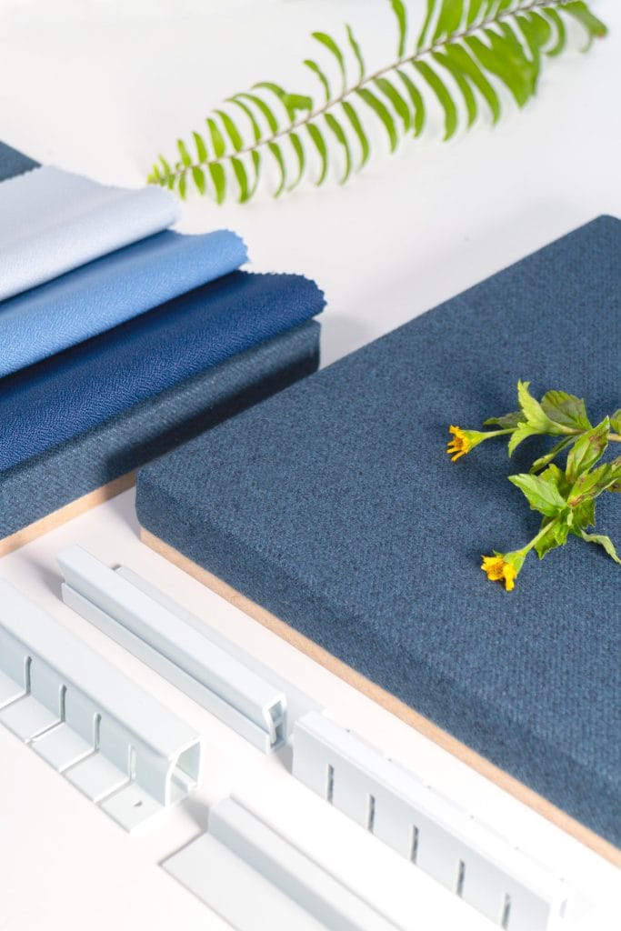 Fabric swatches in shades of blue, textured panels, metal mounting brackets, and green leafy stems are arranged on a white surface, showcasing the versatility of the Magnetic Fabric Track System.