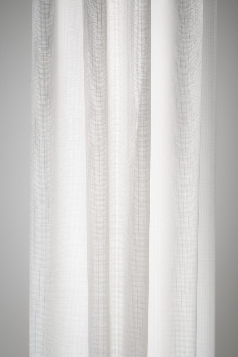 Close-up of white, semi-sheer curtains with a subtle grid pattern—showcasing what makes acoustically transparent fabric—as natural light creates gentle vertical folds and shadows against a neutral background.
