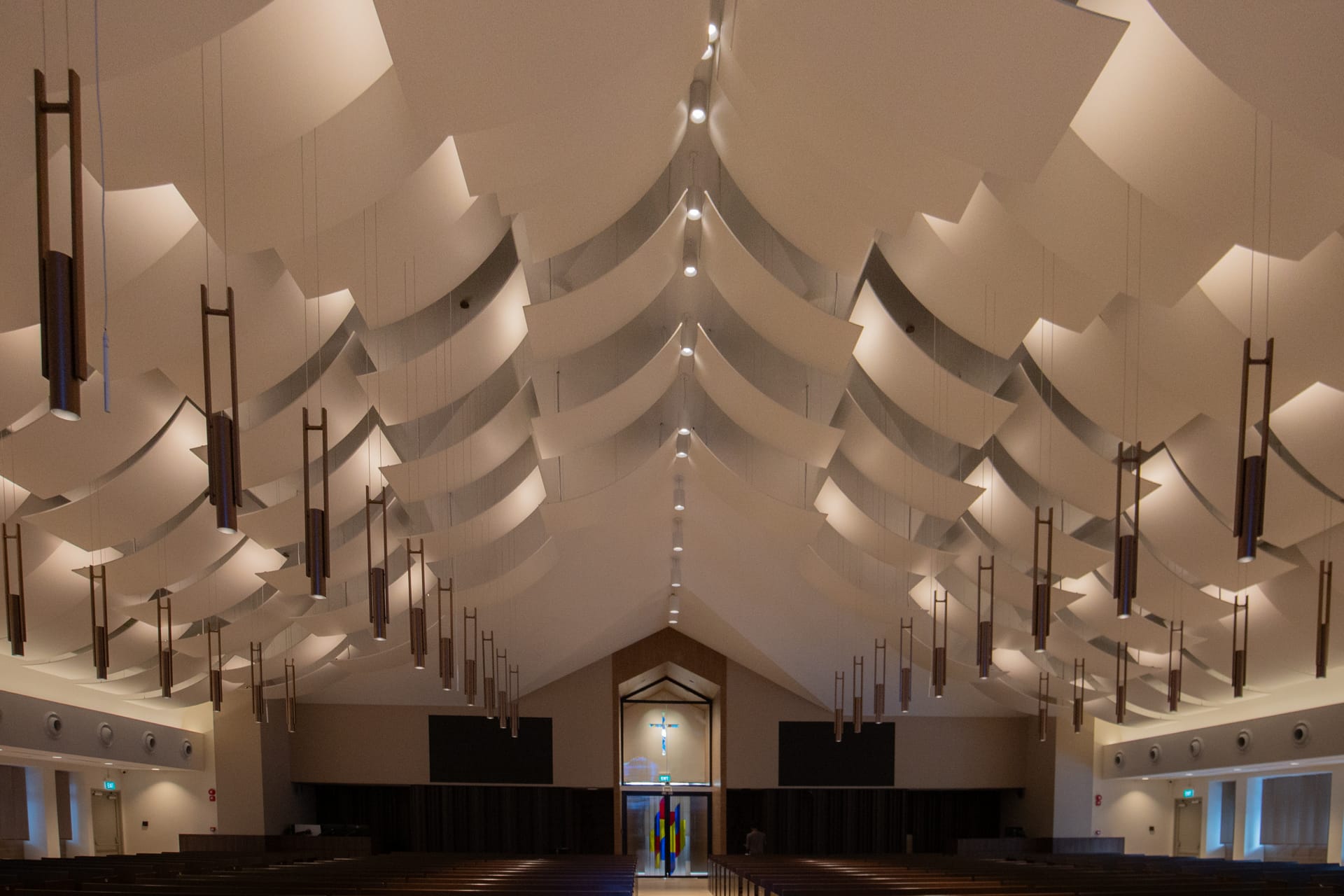 The interior of a modern church features geometric ceiling panels with acoustical fabric for worship spaces, rows of hanging light fixtures, wooden pews, and a large window at the far end displaying a cross and colored glass.