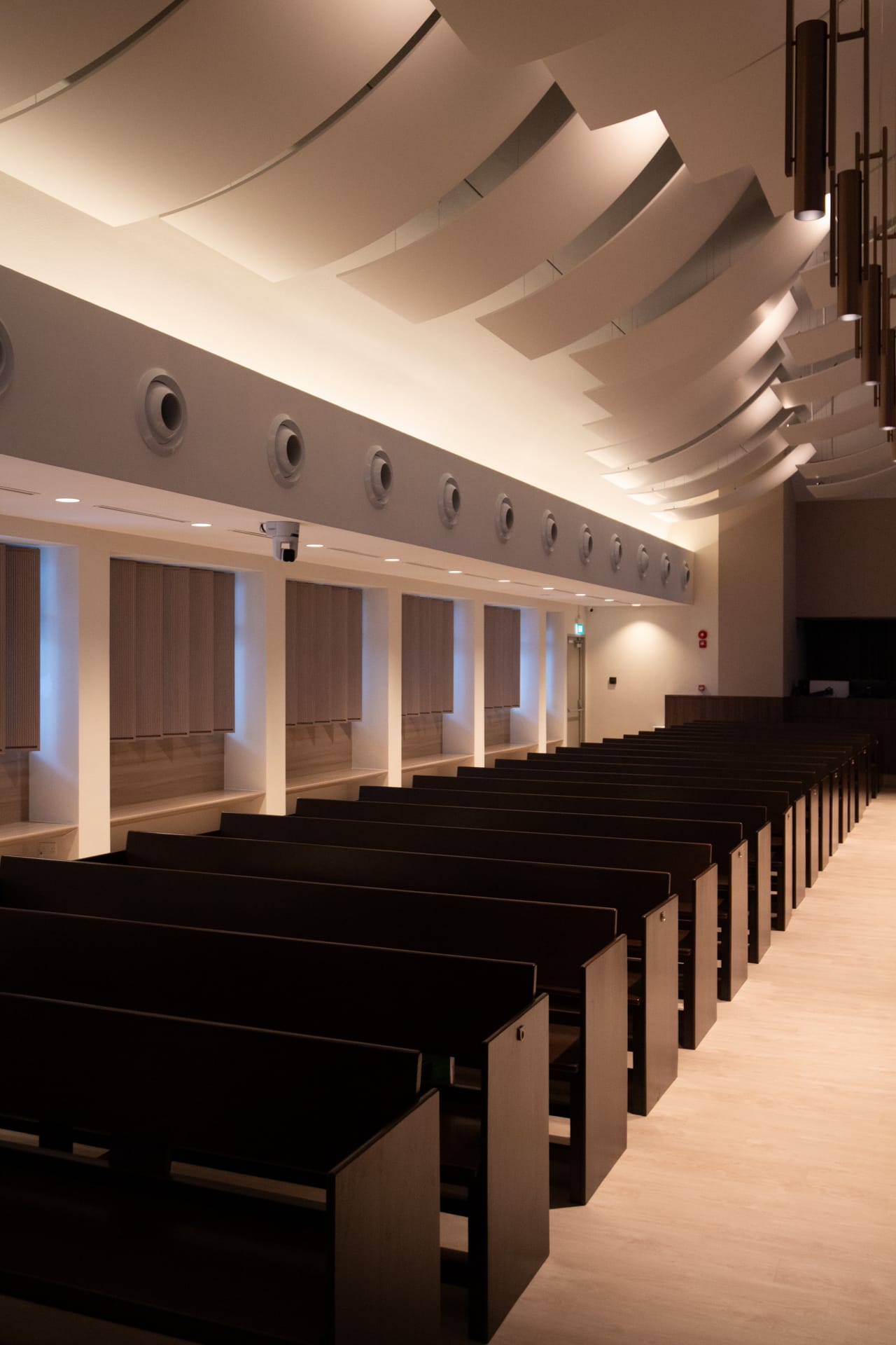 Rows of dark wooden pews in a modern, softly lit chapel with a curved ceiling, circular vents along one wall, and tall windows partially covered by blinds. Acoustical fabric for worship spaces helps enhance the calm and orderly atmosphere.