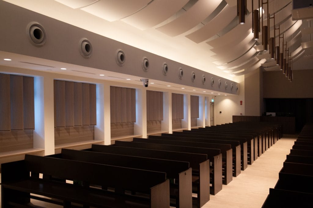 A modern, empty room with wooden benches arranged in rows, beige walls adorned with acoustical fabric for worship spaces, tall windows with blinds, circular vents, and contemporary ceiling lights. The ambiance is calm and softly lit.