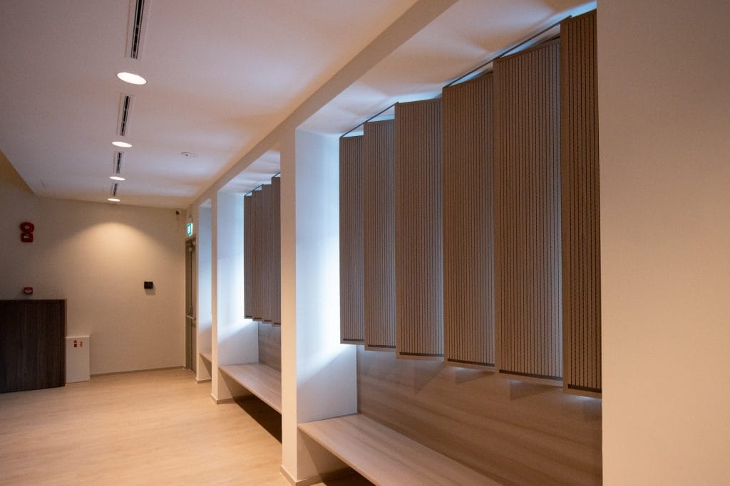 A modern hallway with light wood floors and built-in wooden benches features vertical blinds over tall windows. Neutral walls, ceiling lights, and acoustical fabric for worship spaces add to the clean, minimalist aesthetic.