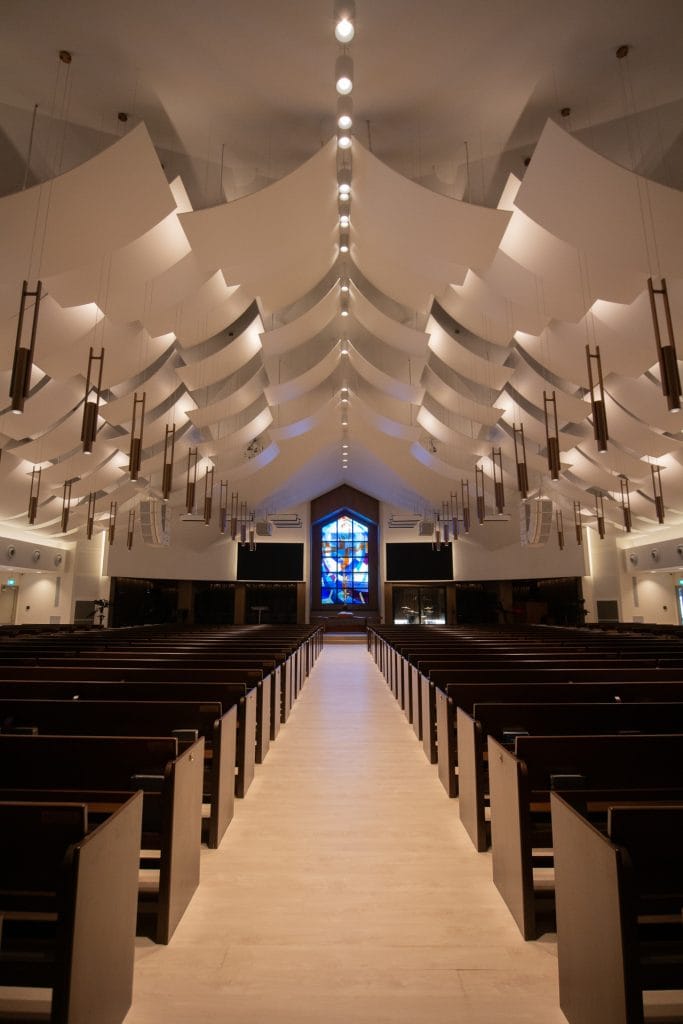 A modern church sanctuary interior with rows of wooden pews, dramatic white ceiling panels enhanced by acoustical fabric for worship spaces, hanging lights, and a colorful stained glass window at the front. The view is from the aisle looking toward the altar.