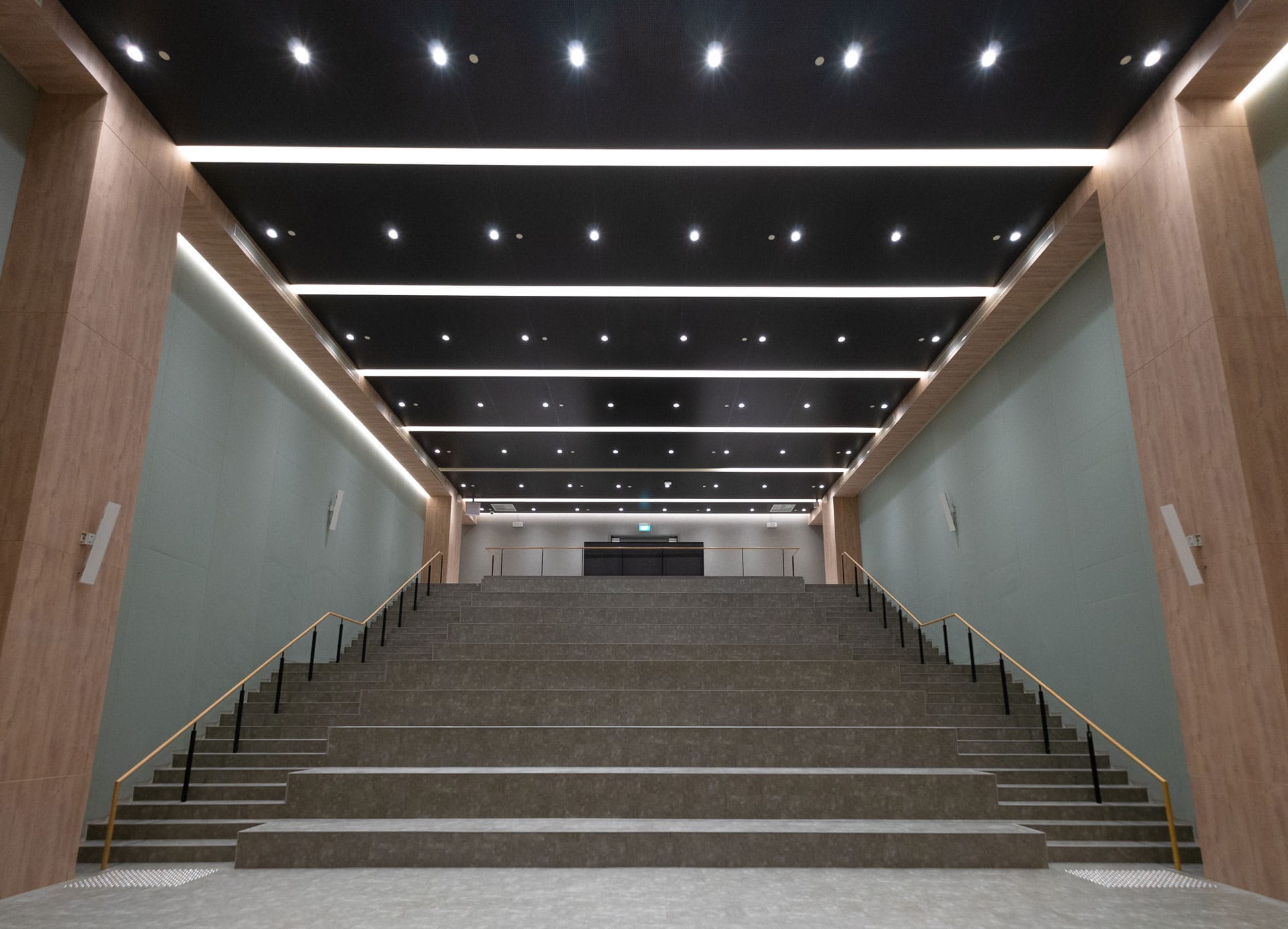 A wide staircase with multiple steps leads up to a landing in a modern interior with high ceilings, wood-paneled walls, black ceiling panels, and rows of bright ceiling lights.