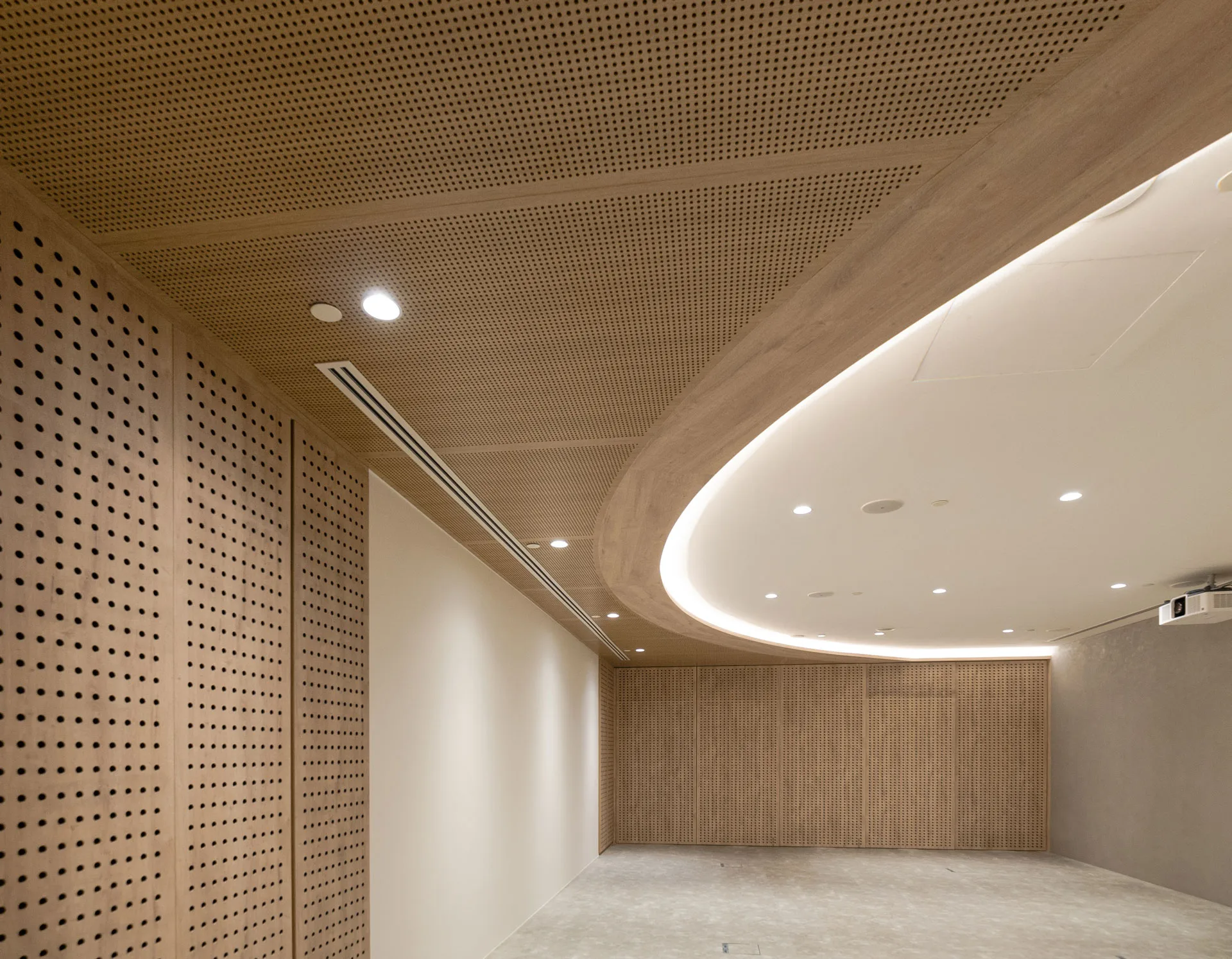 A modern, empty room with light wood-paneled walls and ceiling. The ceiling features recessed lighting and a large, curved illuminated section. The floor is plain, and a projector is mounted near the ceiling.