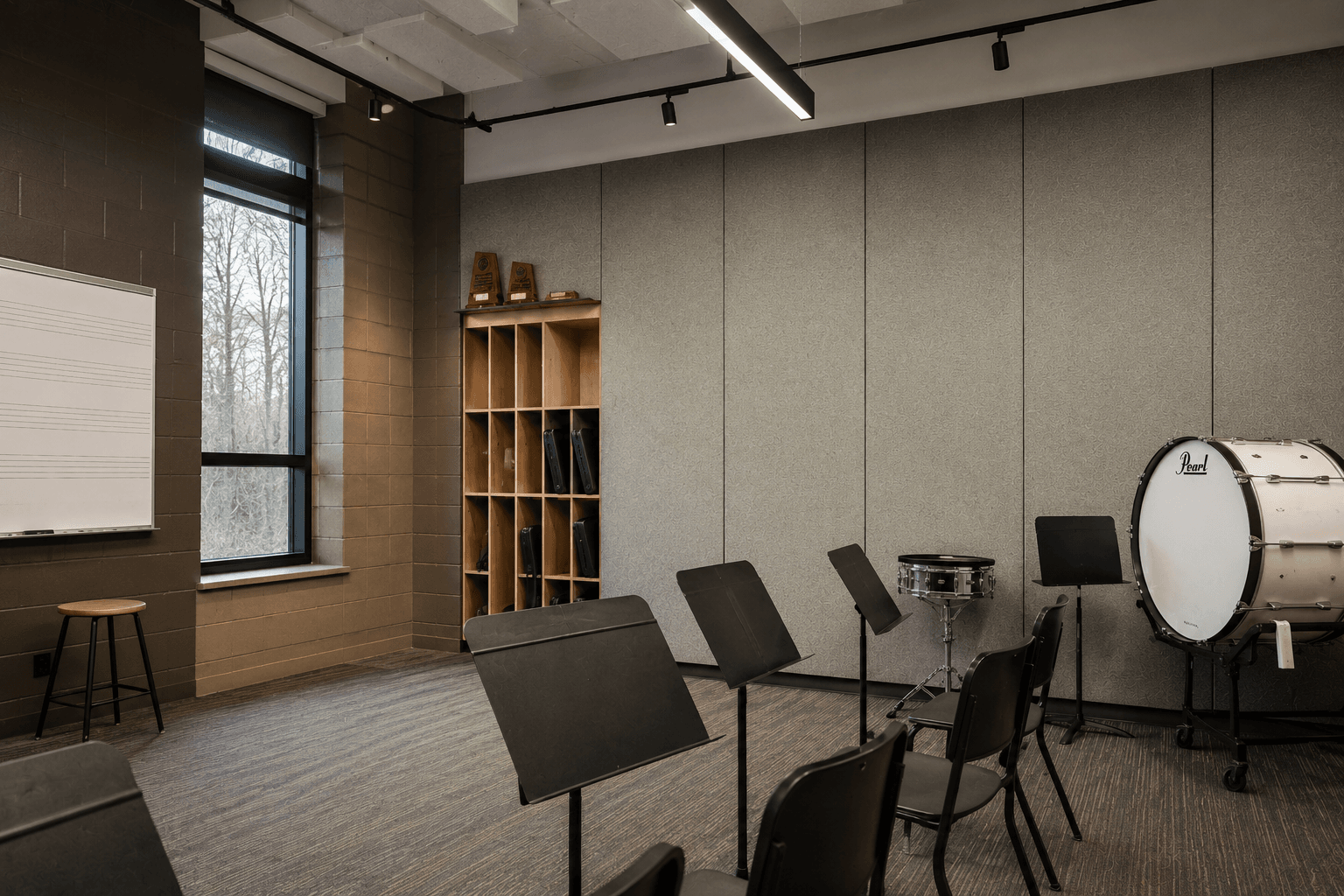 A music classroom with chairs, music stands, a whiteboard with musical staff, a window, shelves holding percussion instruments, and a large bass drum against a wall with acoustic panels.