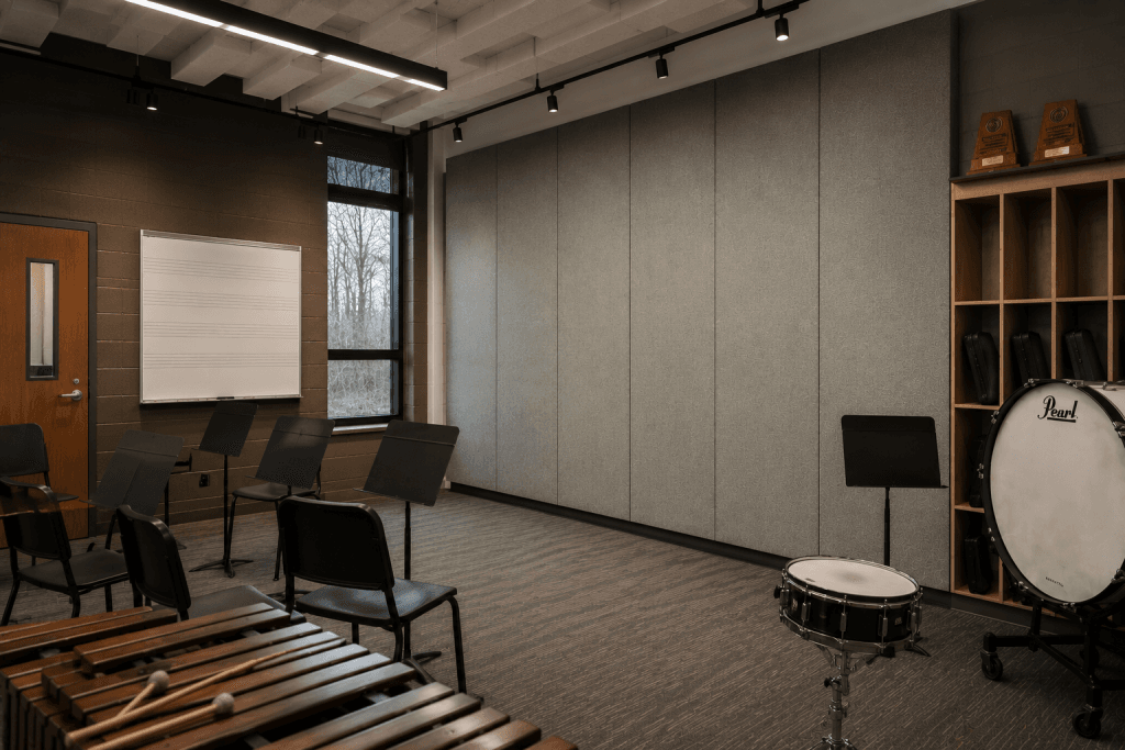 A music classroom with empty chairs and music stands, a whiteboard with musical staff lines, percussion instruments like a xylophone, bass drum, snare drum, and shelves with instrument cases and trophies.