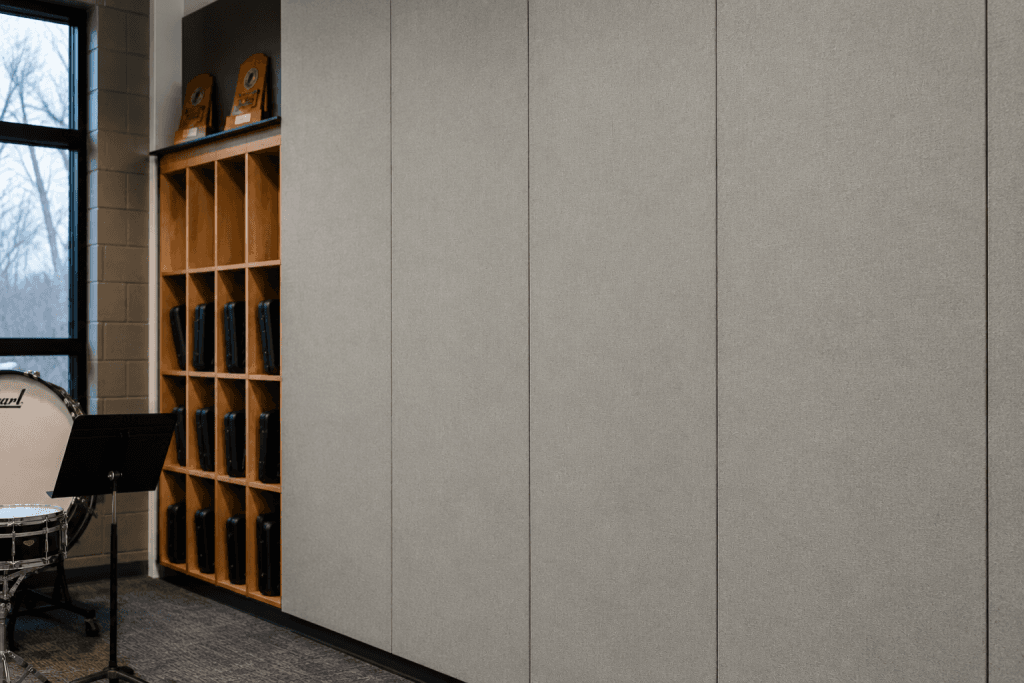 A music room with wooden cubbies holding black instrument cases, a large drum, a music stand, and award plaques on top of the cubbies; grey acoustic panels cover the adjacent wall.