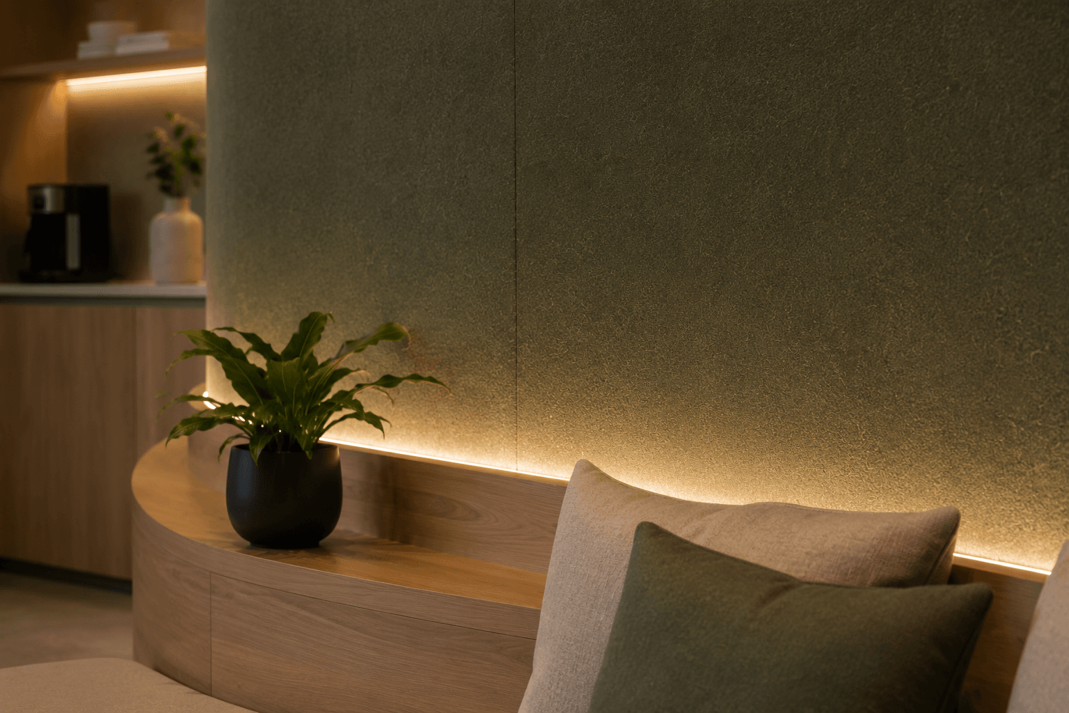 A modern lounge area with a green textured wall, warm underlighting, a wooden bench, a potted plant in a black pot, and neutral-toned cushions on a beige sofa. A kitchen counter is visible in the softly lit background.