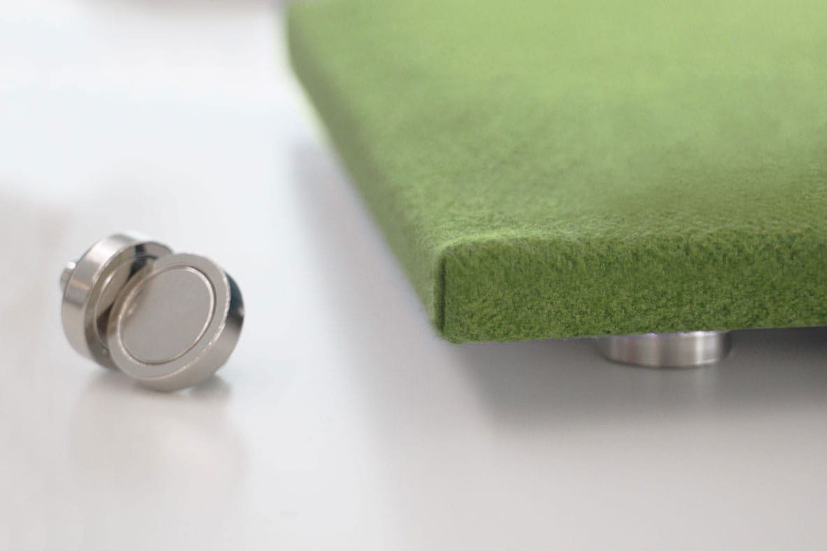 Close-up of a green, fabric-covered panel with metallic feet on a white surface, part of a Magnetic Fabric Track System. A small, round metal object lies nearby. The image has a minimal, modern aesthetic.