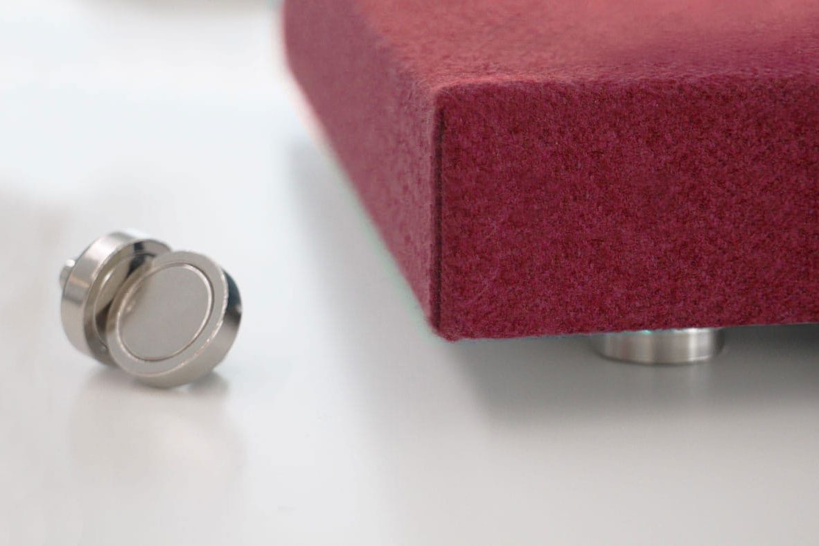 A close-up of a magenta Magnetic Fabric Track System rectangular object with metal feet, resting on a white surface. A small, round, silver metal piece lies nearby.