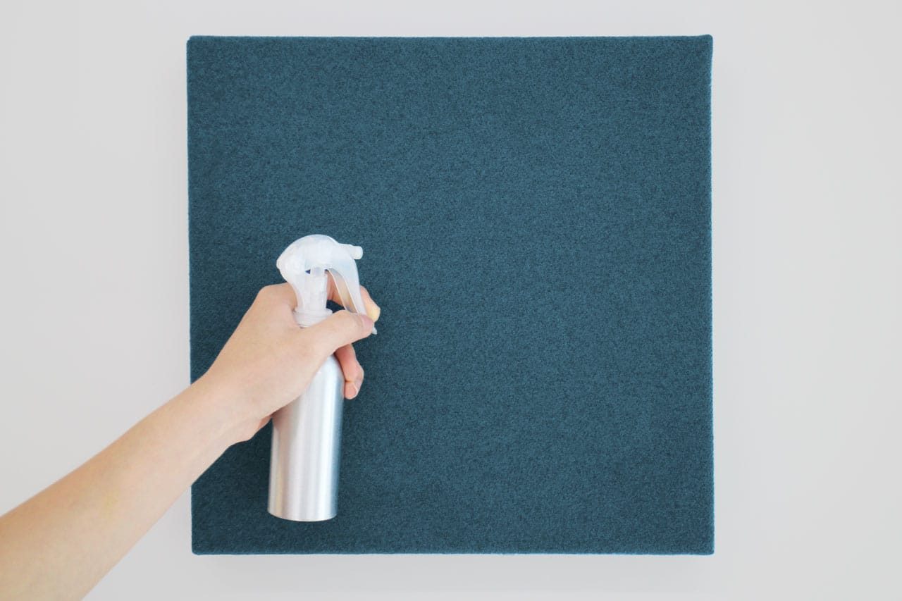A hand holding a silver spray bottle is aimed at the center of a square, teal-colored felt board mounted on a white wall, part of the Fabrix™ Acoustic System Warranty.