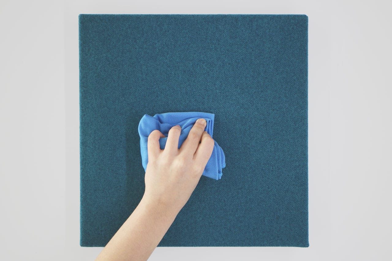 A hand wipes a blue fabric-covered square panel with a blue cloth against a white background, showcasing the easy maintenance of the Fabrix™ Acoustic System Warranty.