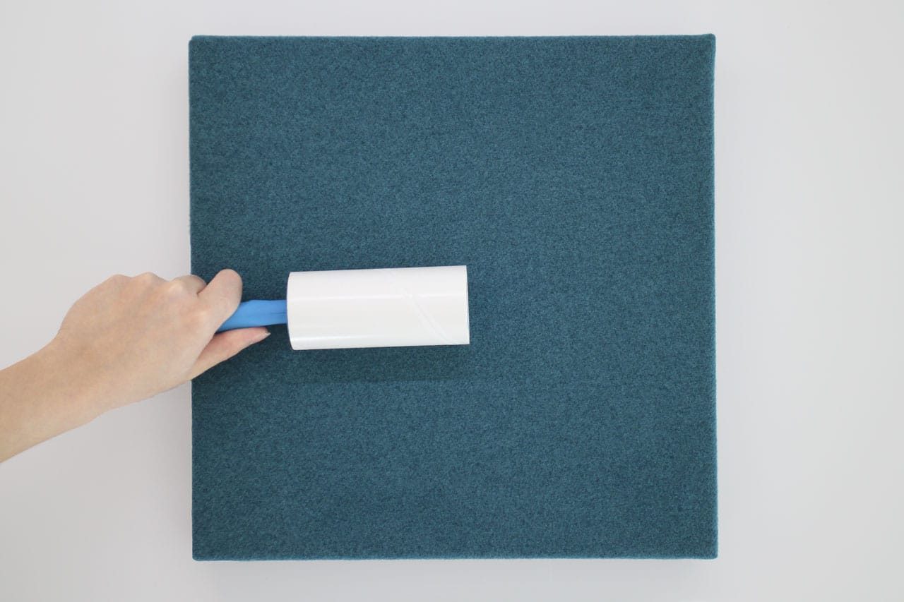 A hand uses a lint roller to clean a square piece of dark teal fabric from the Fabrix™ Acoustic System, highlighting its care and quality assurance under the Fabrix™ Acoustic System Warranty, against a white background.
