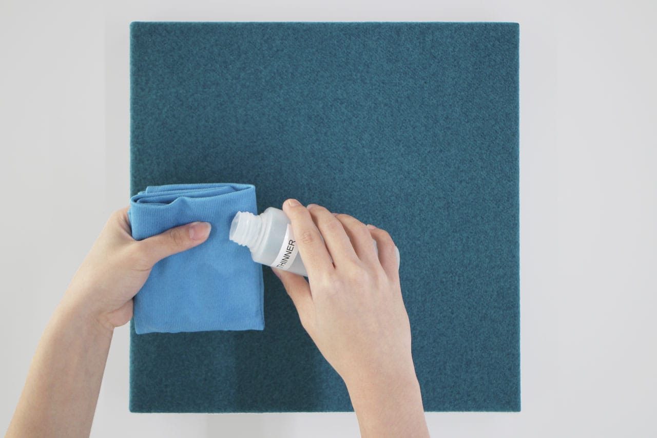 A person pours cleaner onto a folded blue cloth, preparing to clean a teal square fabric panel on a white surface as part of Fabrix™ Acoustic System Warranty care.