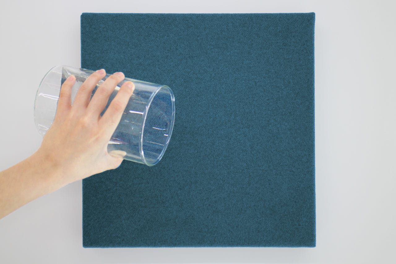 A hand tilts a clear glass cup as if about to pour water onto a square, flat, dark teal fabric surface—part of the Fabrix™ Acoustic System Warranty—set against a white background.