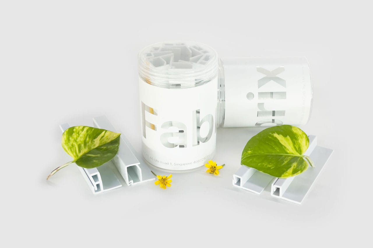 Two white cylindrical containers labeled Fabrix™ Sustainable Acoustic Fabric and two leafy green plants with yellow flowers are displayed on a white background alongside white plastic or metal brackets.