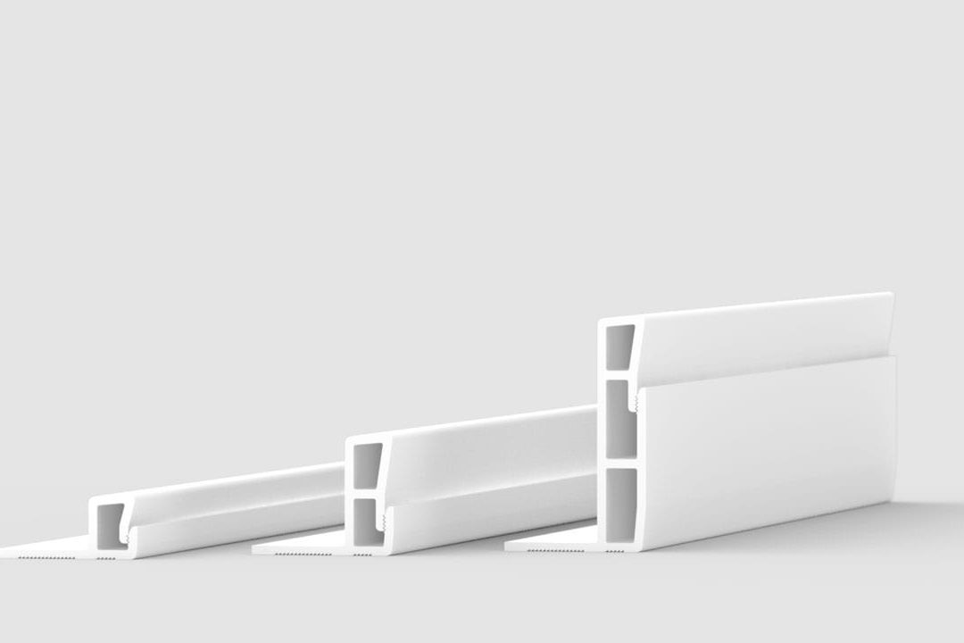 Three white plastic extrusion profiles are arranged on a plain, light gray background. Each profile has a rectangular, hollow structure with different heights and thicknesses, positioned side by side in a diagonal row.