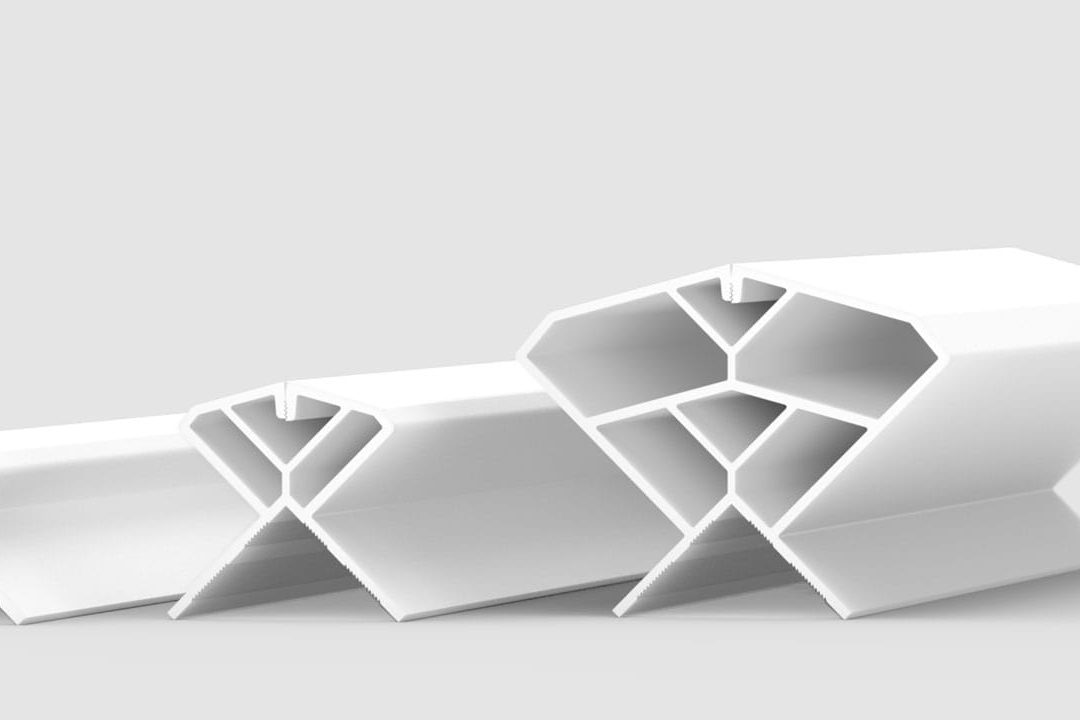 Three white, geometric plastic or metal extrusion profiles of varying sizes are arranged in a row on a plain light background. Each profile has multiple internal chambers and angled surfaces, resembling an abstract honeycomb shape.