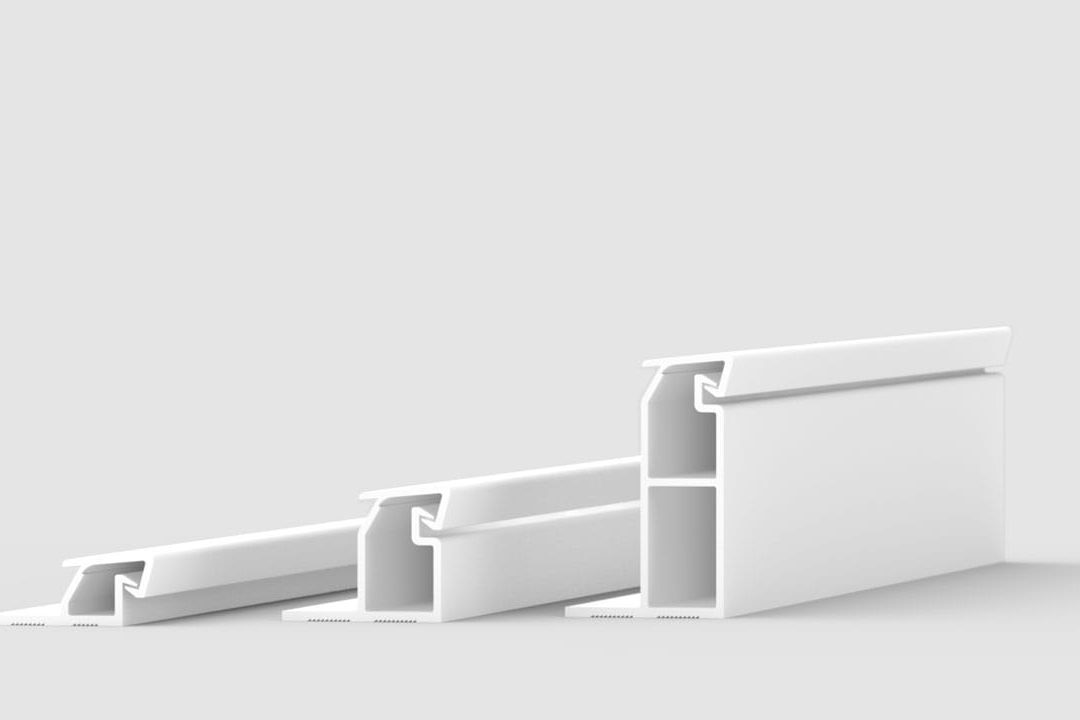 Three white, elongated plastic or metal profiles of varying sizes are arranged in a row on a light gray background, each featuring a hollow rectangular and angular cross-section.